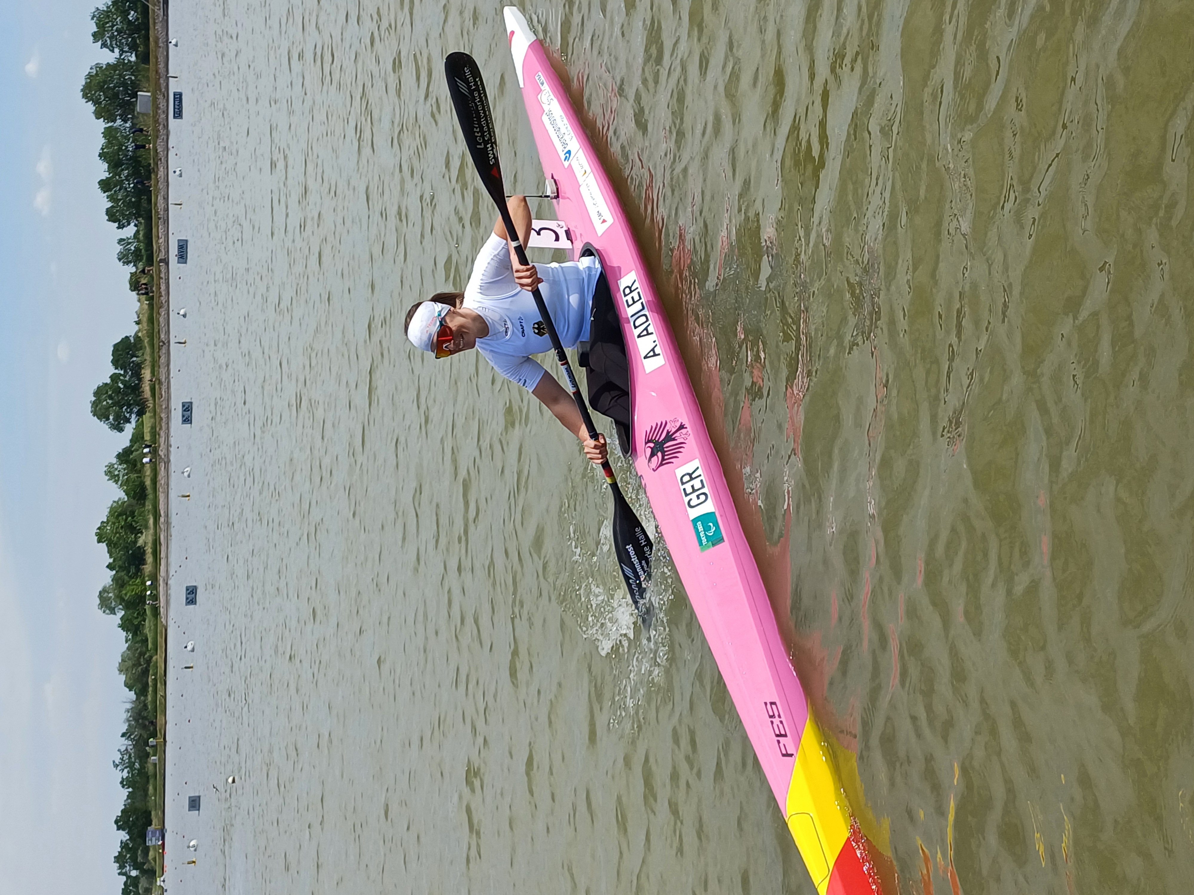 Anja in ihrem rosafarbenen Kanu mit einem Doppelpaddel und fährt auf einem See. Sie lächelt und trägt eine Sonnenbrille sowie ein weißes Kopftuch.
