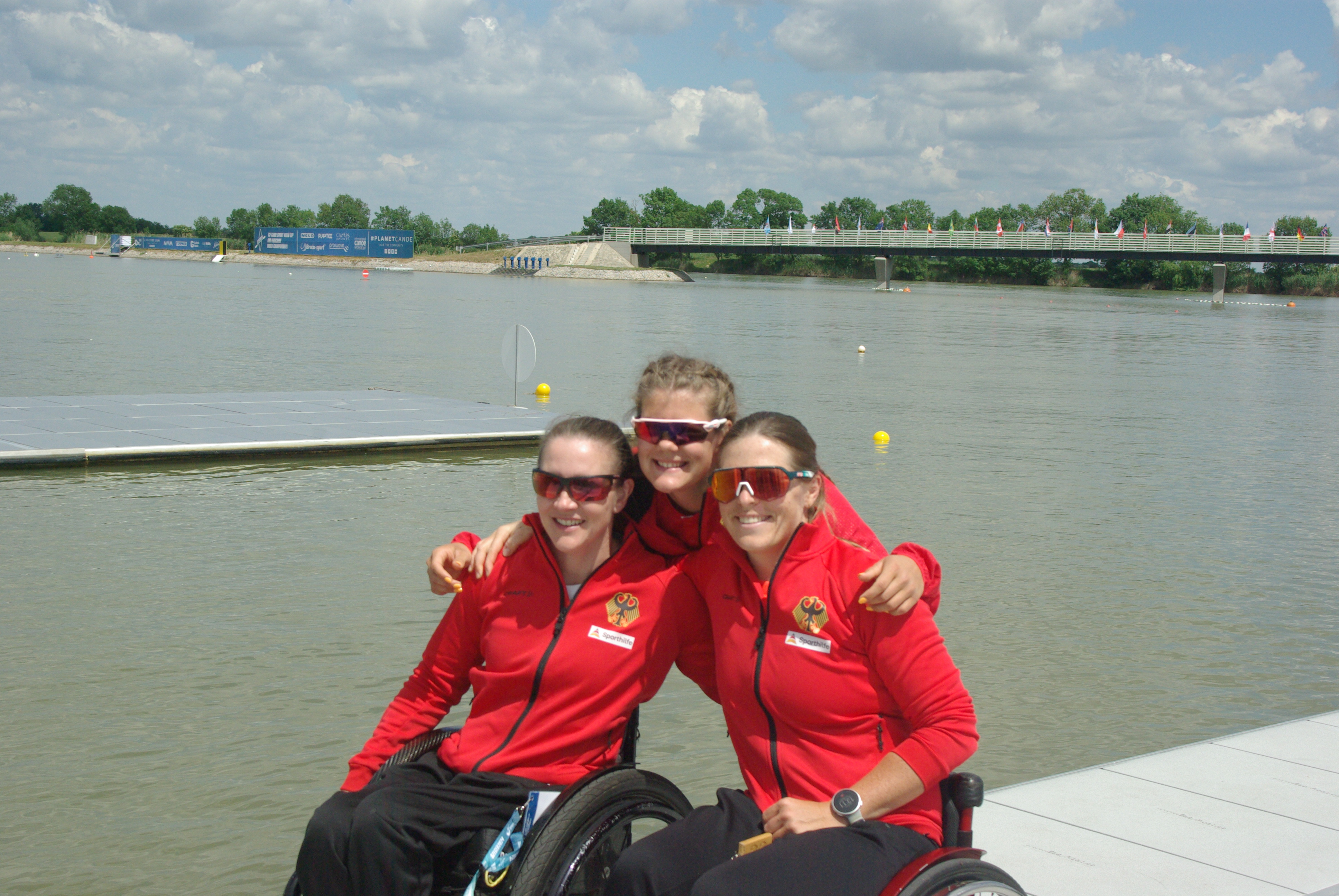 Drei Frauen mit Sonnenbrillen und roten Jacken lächeln in die Kamera. Zwei von ihnen sitzen im Rollstuhl, eine steht (gebückt) in der Mitte. Im Hintergrund ist ein See zu erkennen.