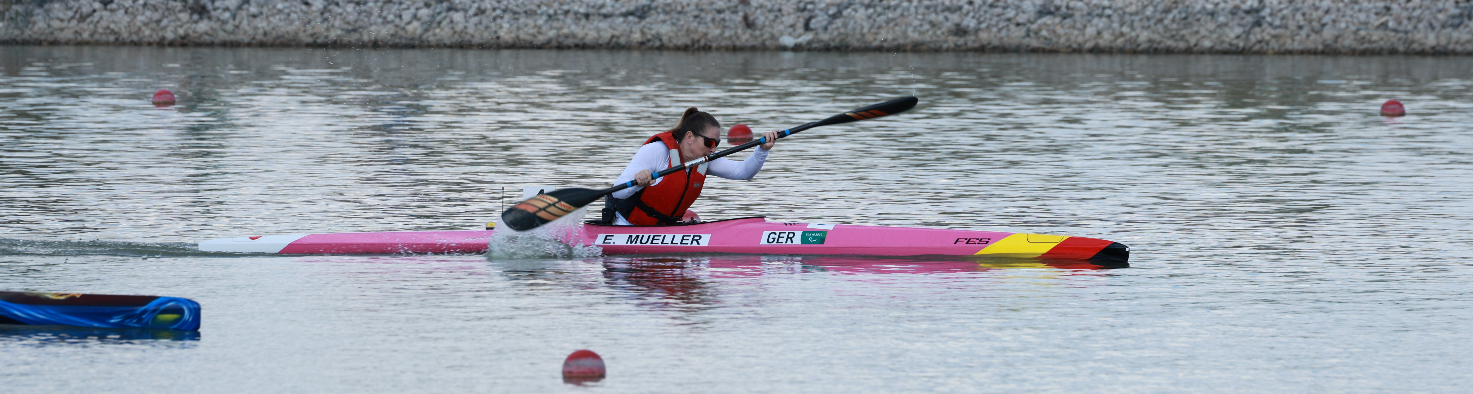 Para-Kanutin Edina Müller in ihrem rosafarbenen Kanu bei voller Fahrt. Sie trägt eine Sonnenbrille und das rote Trikot der Nationalmannschaft.