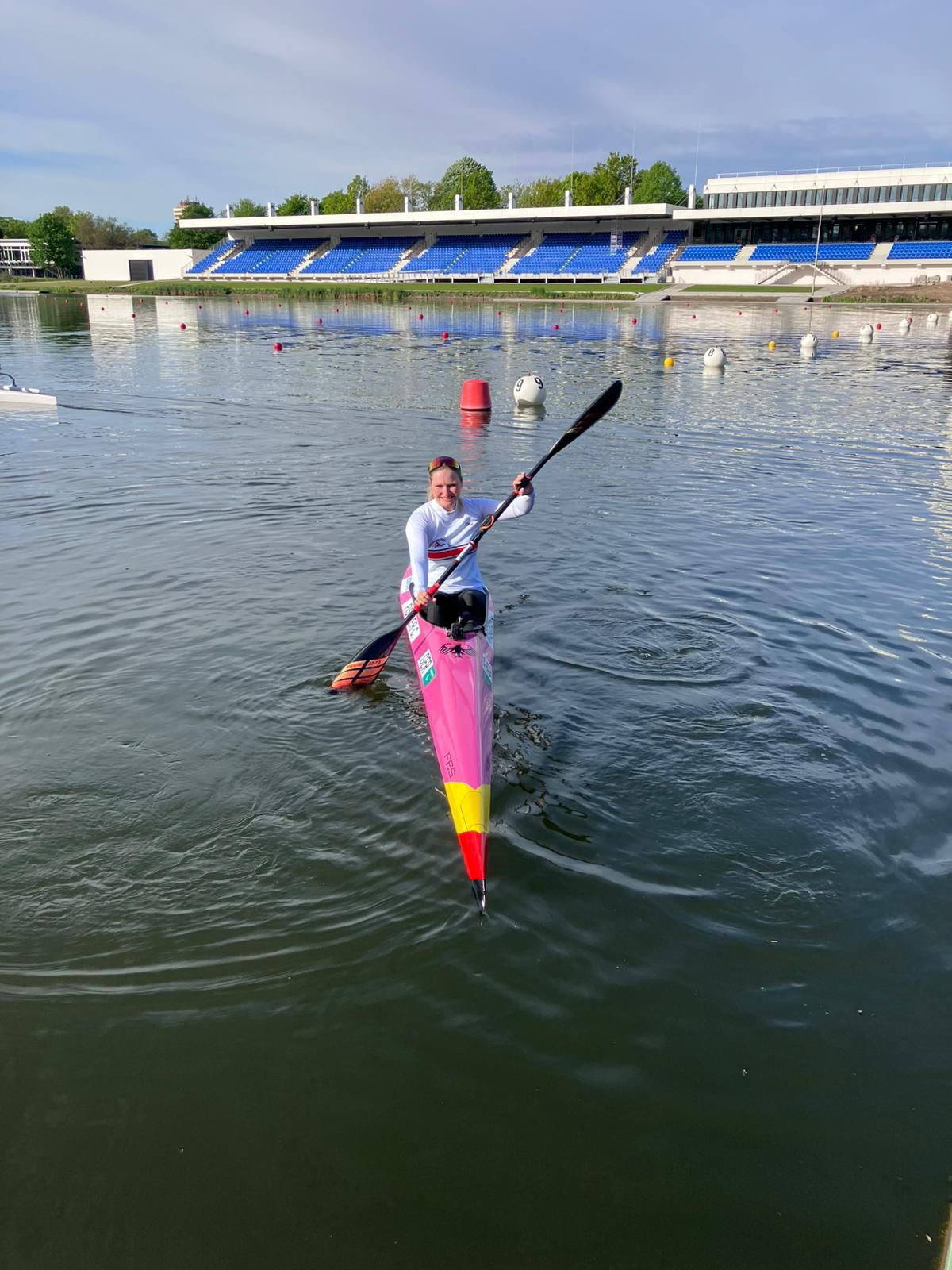 Kanufahrerin Claudia Fischer in einem pink-gelben Rennkajak auf einer Regattastrecke mit Tribüne im Hintergrund.