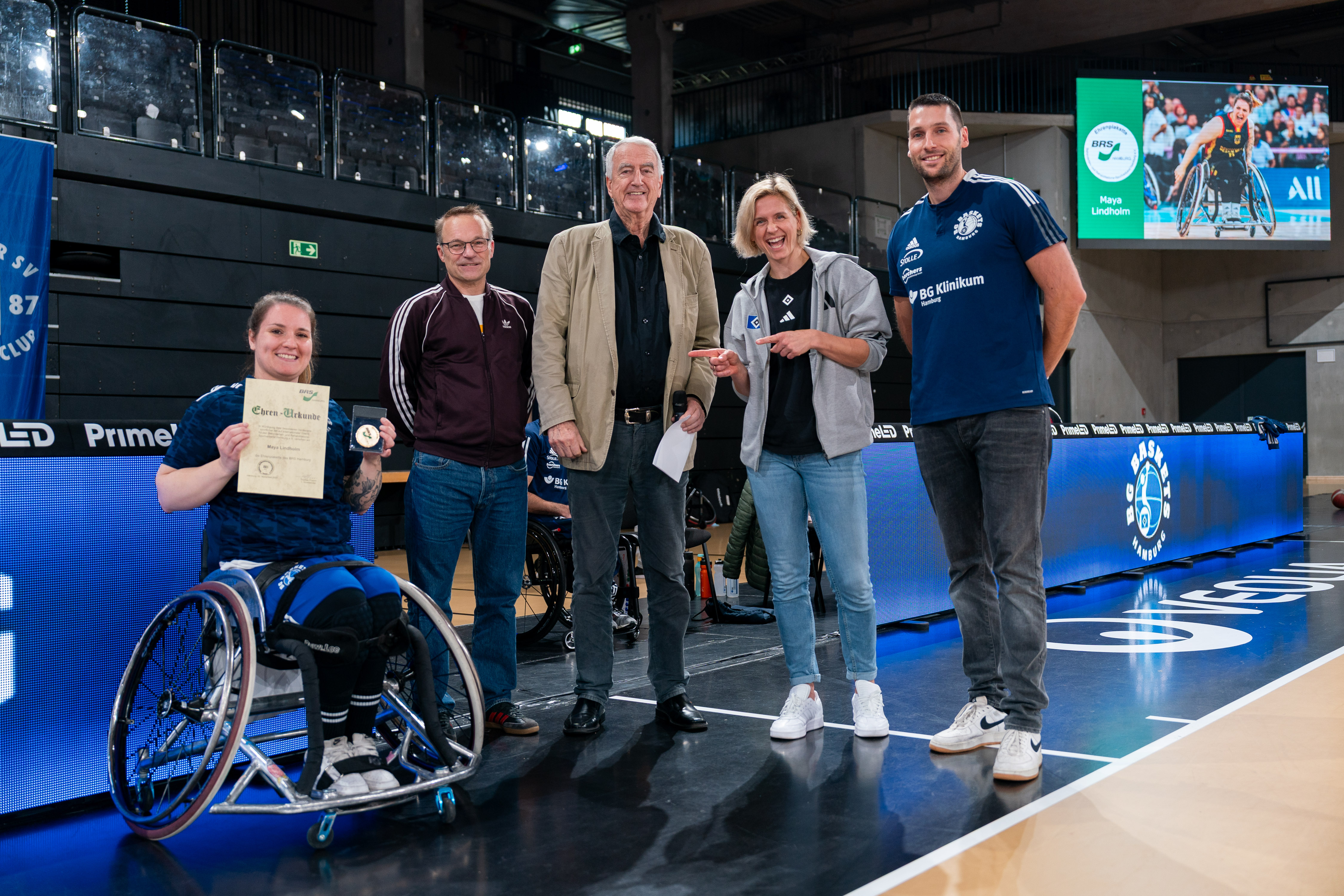 Maya sitzt in ihrem Rollstuhl und lächelt in die Kamera. Dabei hält sie die Plakette und die Urkunde des BRSH hoch.