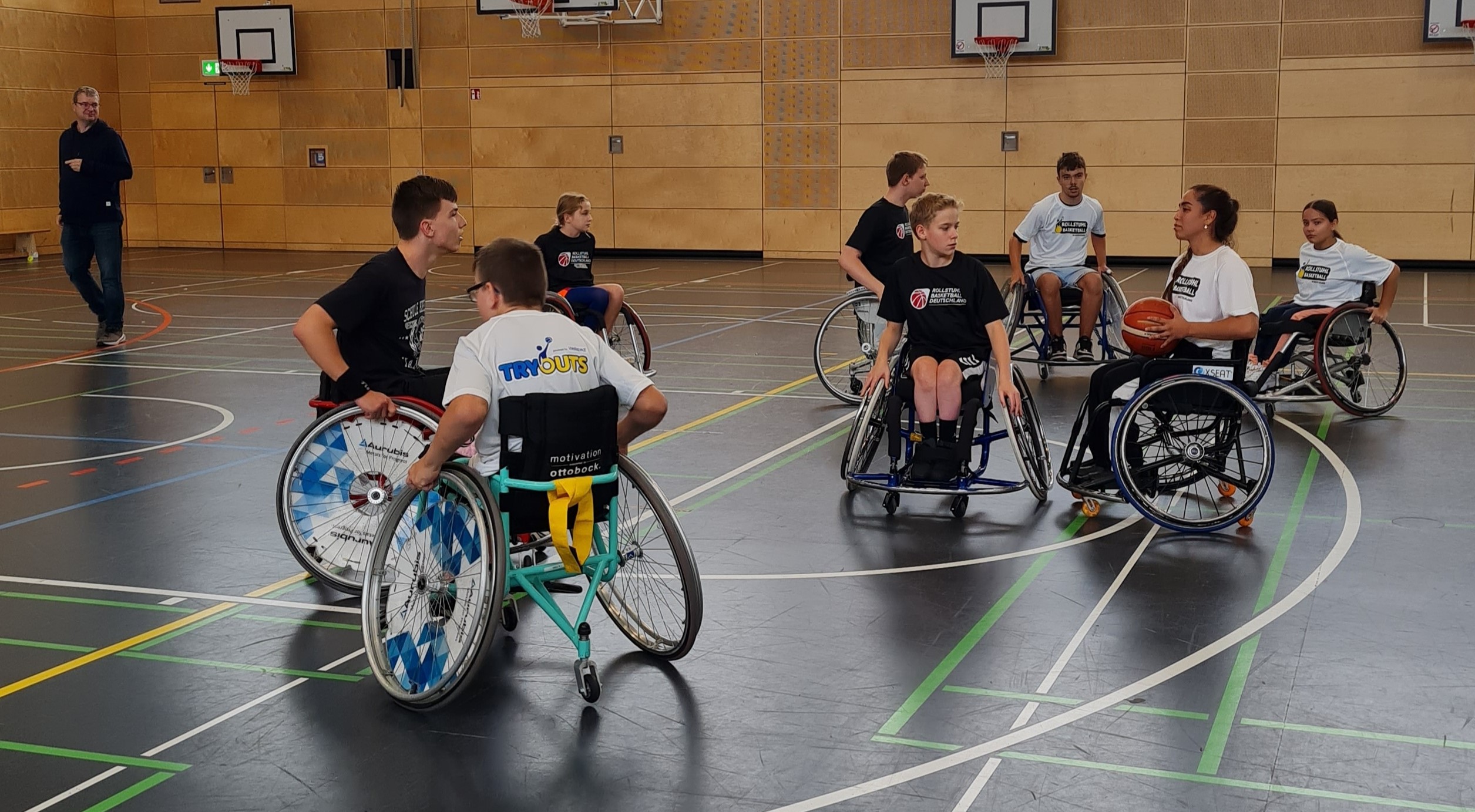 Mehrer Rollstuhlfahrenden in eine Halle mit dunklem Boden. Eine Teilnehmerin hat einen Basketball in den Händen.