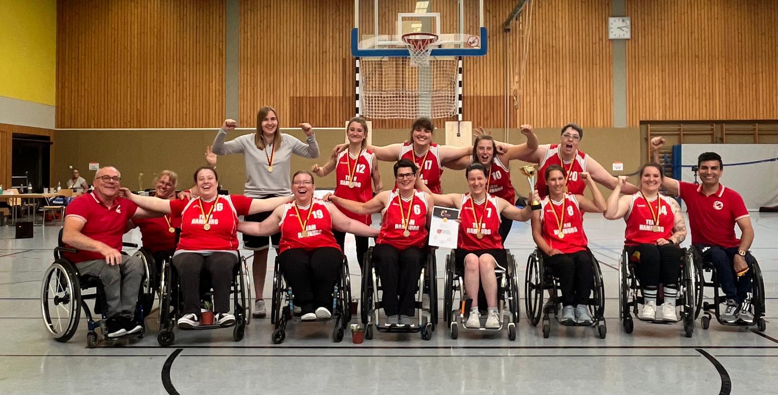 Das Team der Rollstuhbasketball-Nationalmannschaft mit Trainer:innen Team. Alle tragen das National-Trikot, halten sich in den Armen und lächeln.