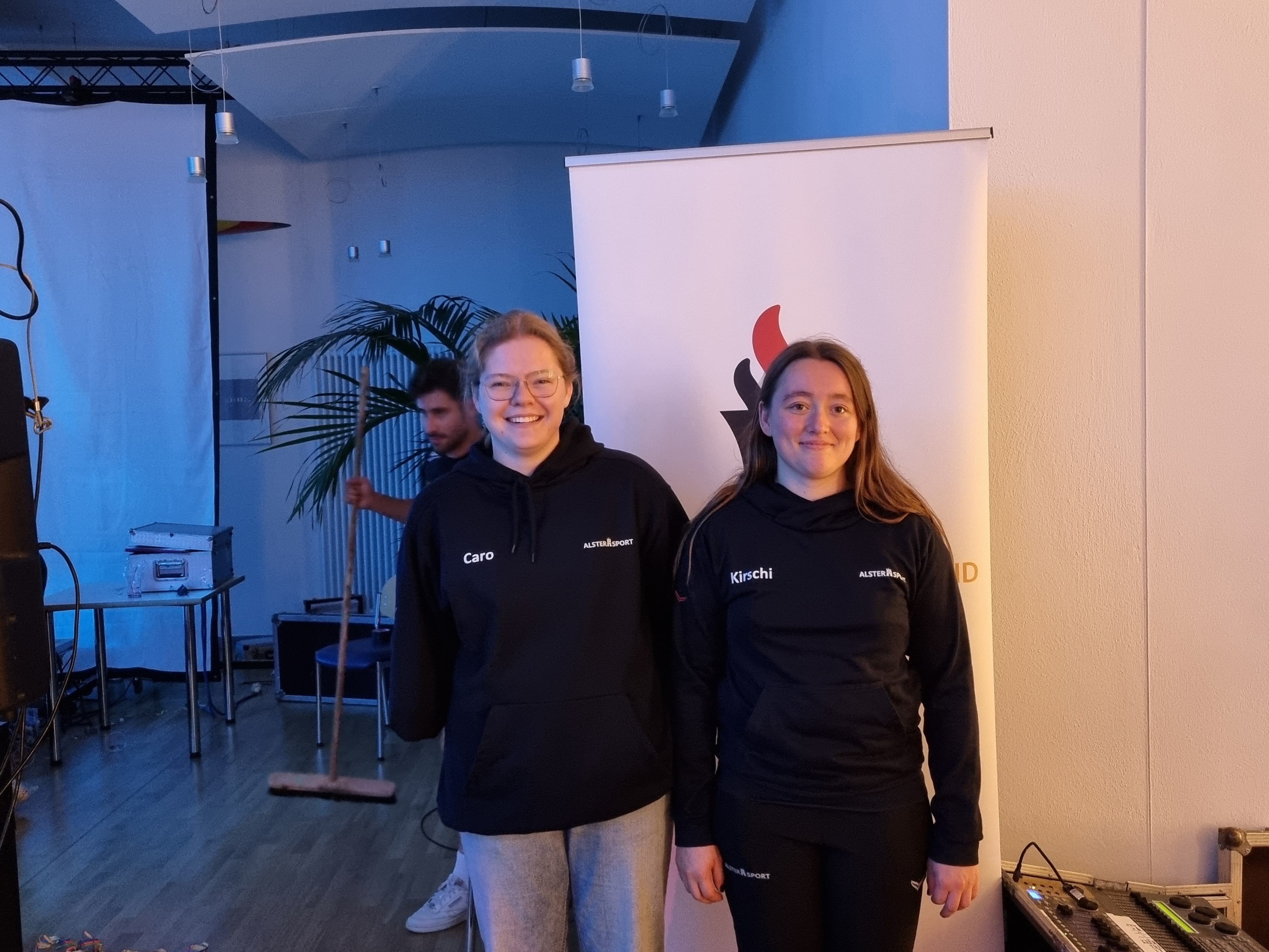 Zwei junge Frauen stehen nebeneinander vor einem Aufsteller mit Vereinslogo. Beide tragen dunkelblaue Trainingsjacken, links ‚Caro‘, rechts ‚Kirschi‘. Sie schauen in die Kamera und lächeln.